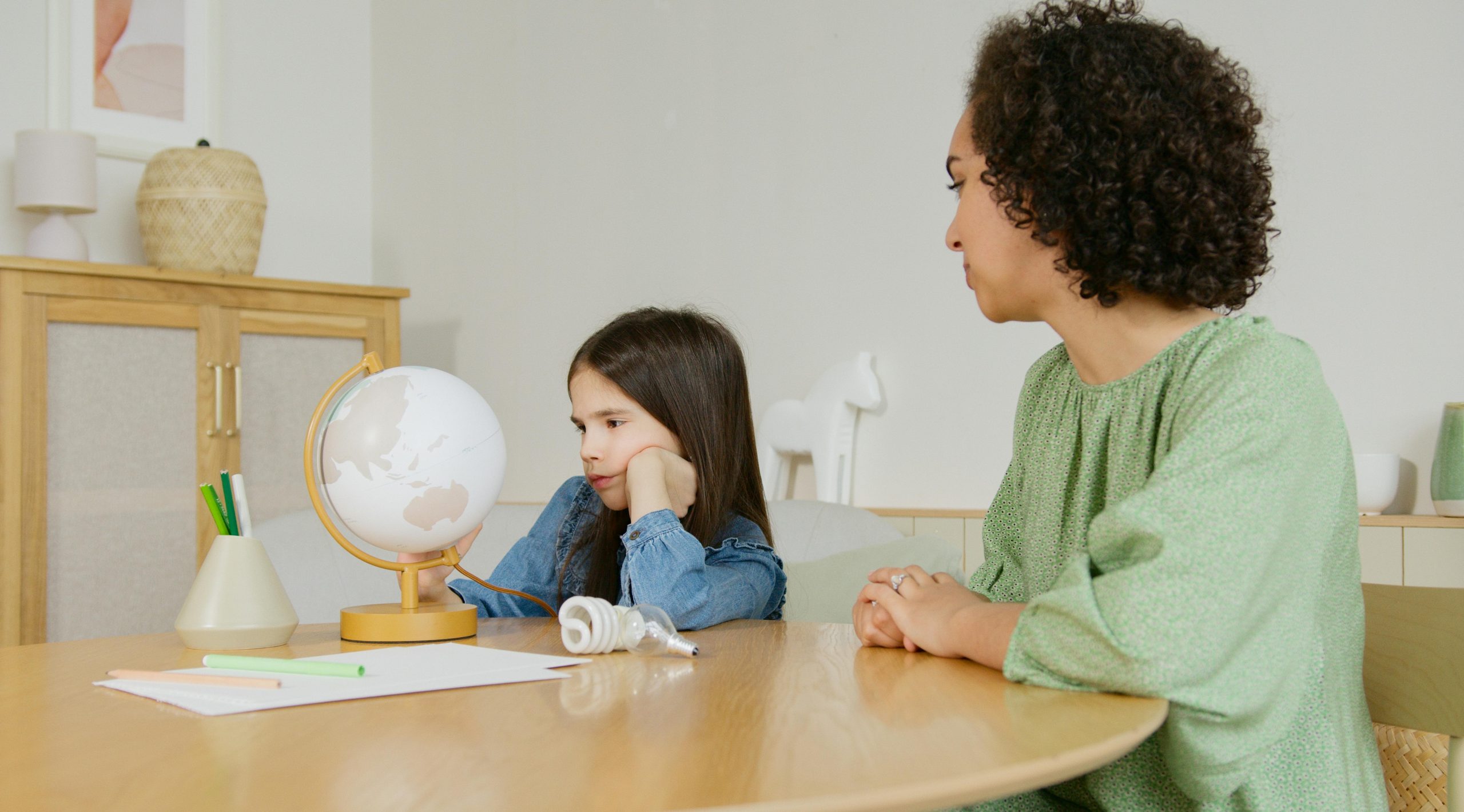 A child looks at a globe on the table as her mother watches on