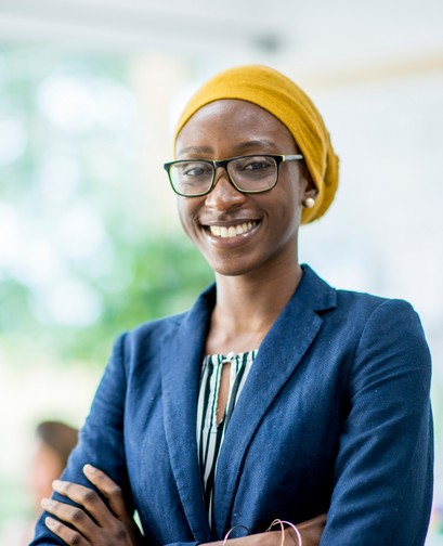 A woman of colour in a yellow headscarf and glasses has her arms folded. She is smiling at the camera and wearing a blue blazer.