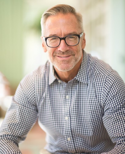 A professional-looking man wearing glasses and a long sleeve button down shirt is smiling at the camera.