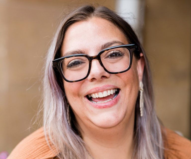 A woman with grey-purple hair and glasses is smiling at the camera.