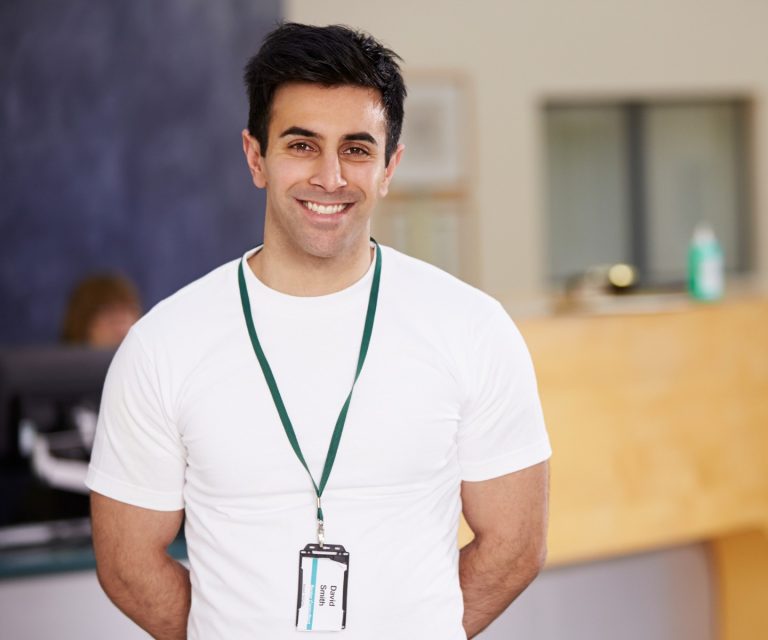 A young man is wearing a white tee shirt and a lanyard. He has his arms behind his back and he is smiling at the camera. He is standing in the reception area of a professional workplace.