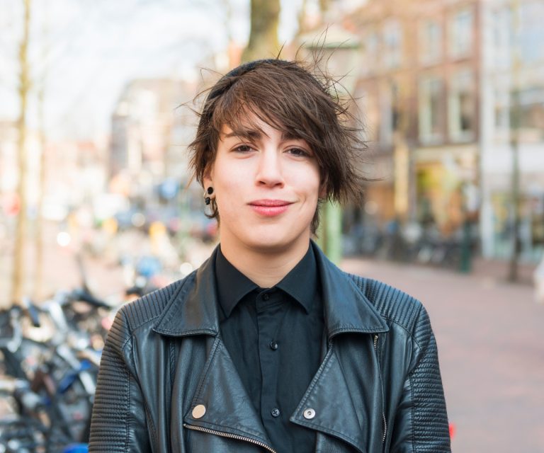 A non-binary person with short spiked hair and a leather jacket is standing in front of a quiet street with parked motorcycles. They are smiling at the camera.