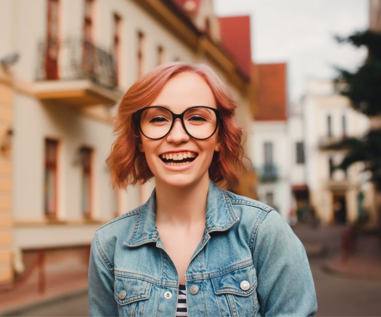 A young woman with red hair and glasses is walking down a suburban street, smiling at the camera.