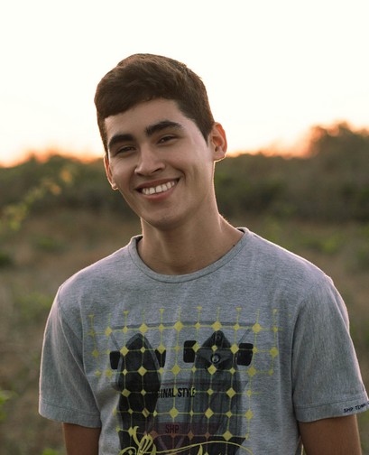 A young man stands in a field. He has short hair and a tee shirt, and he is smiling at the camera.