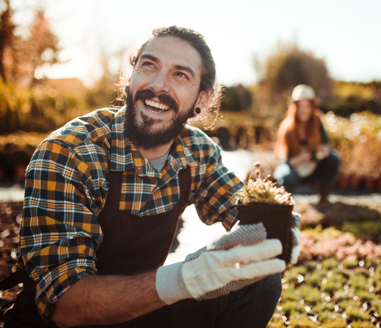 A young man with a beard is working in a nursery. He is crouching and holding a seedling while smiling.