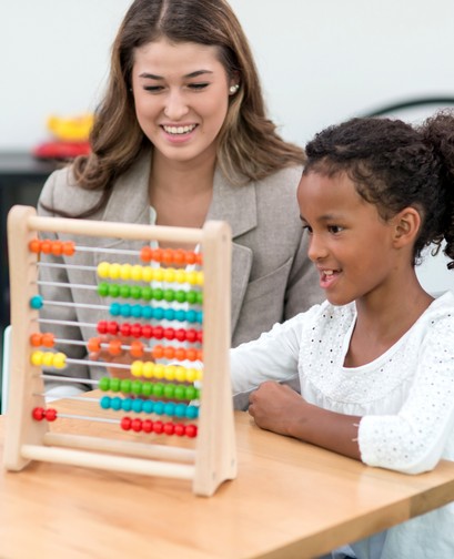 A female teacher with long hair sits with a young female student of colour. They are looking at an abacus and smiling.