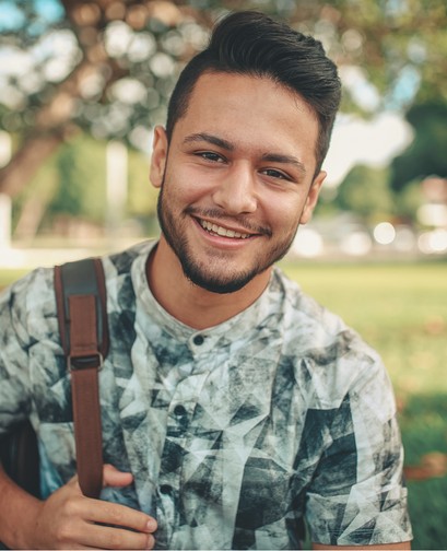 A young man is holding a backpack over his shoulder and smiling at the camera.