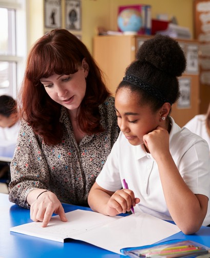 A female teacher with long red hair is pointing at an open notebook that sits in front of her student. Her student is a young girl of colour with her hair in a bun. She is holding a pen and looking attentively at her teacher.