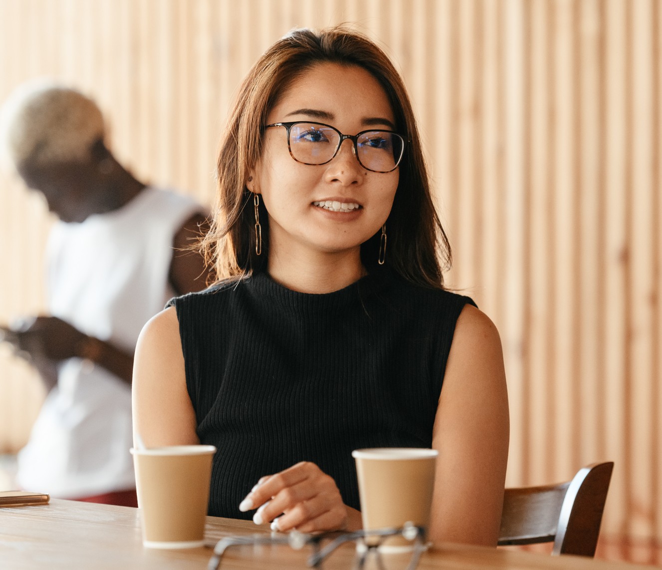 A young Asian woman is dressed professionally and is sitting at a cafe. She has two coffee cups in front of her and she is wearing glasses. She is speaking to someone.