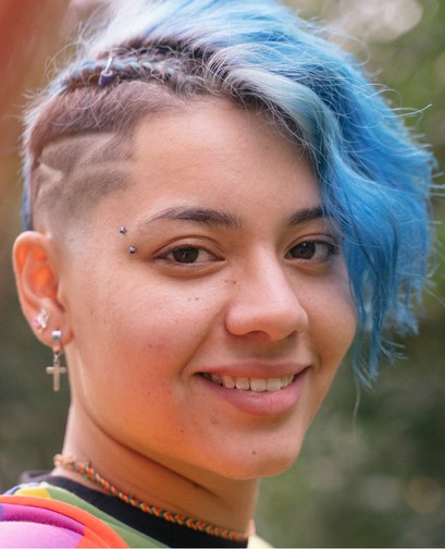 A young woman with blue shaved hair and facial piercings is smiling at the camera.