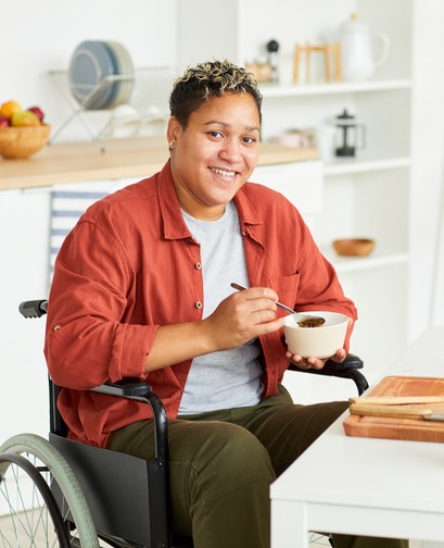A non-binary person with short dark hair is in a wheelchair, sitting in their kitchen. They are eating a bowl of cereal and looking at the camera smiling.