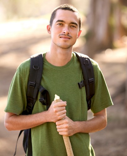 A young man in a tee shirt and backpack is hiking, and holding a hiking pole. He is looking at the camera and smiling.