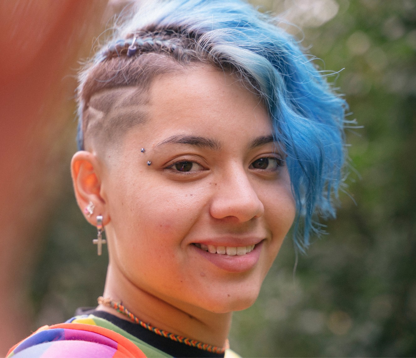 A young woman with blue shaved hair and facial piercings is smiling at the camera.