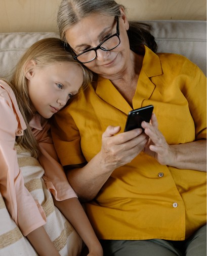 A grandmother and young granddaughter are sitting on a couch. The granddaughter is laying her head on her grandmother's shoulder. The grandmother is holding an iPhone and showing the granddaughter. They are both smiling at the phone.