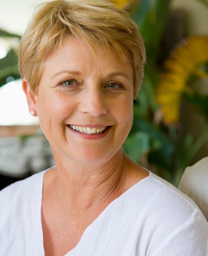 A middle aged woman with short blonde hair is wearing a white blouse. She is smiling at the camera.
