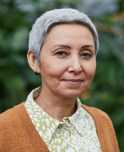 An older woman with short grey hair is looking at the camera, smiling softly. She is standing outside and wearing an orange cardigan and yellow blouse.