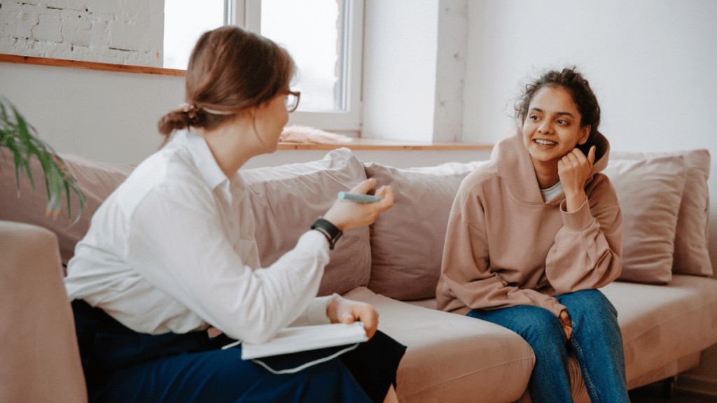 A teenage girl sits on a couch smiling. An older, professional woman sits in front of her, holding a notebook and pen. They are having a conversation.