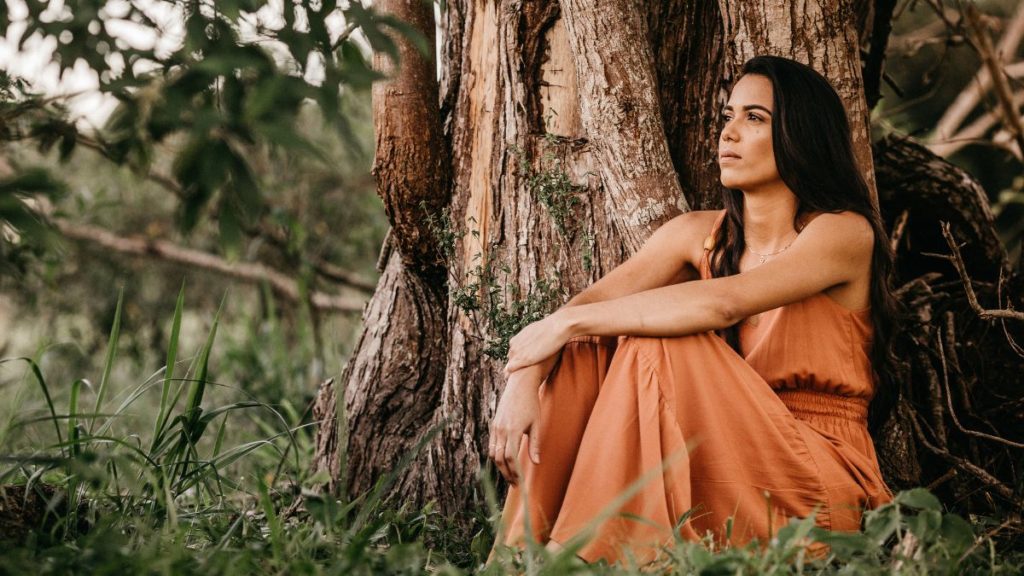 A woman wearing a flowing long dress sits in front of a tree, looking off into the distance.