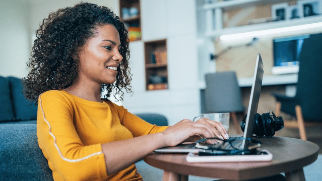 A woman of colour sits at her laptop in her living room. She is typing and smiling at her laptop.