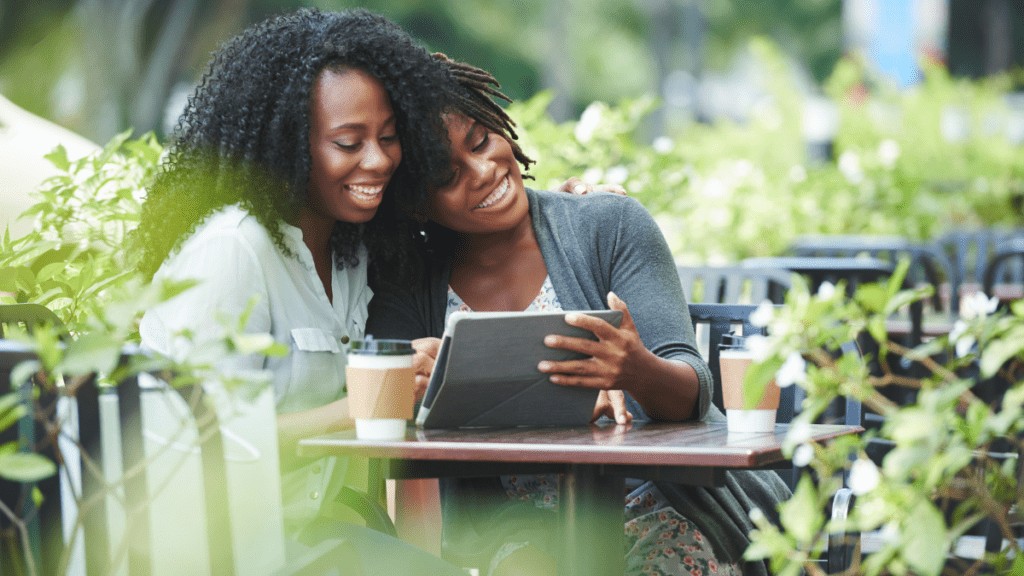 Two women of colour sit at a cafe, using an iPad. One woman leans her head on the shoulder of the other woman. Both are smiling at the iPad.