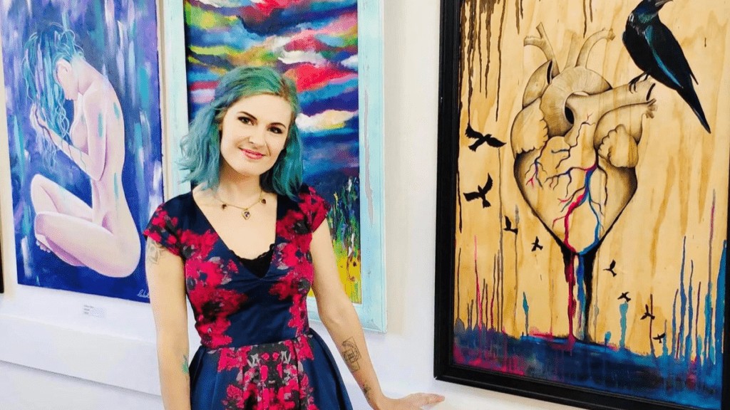 A woman wearing a black and red dress with blue and green hair is standing in front of a series of paintings. She is looking at the camera and smiling.