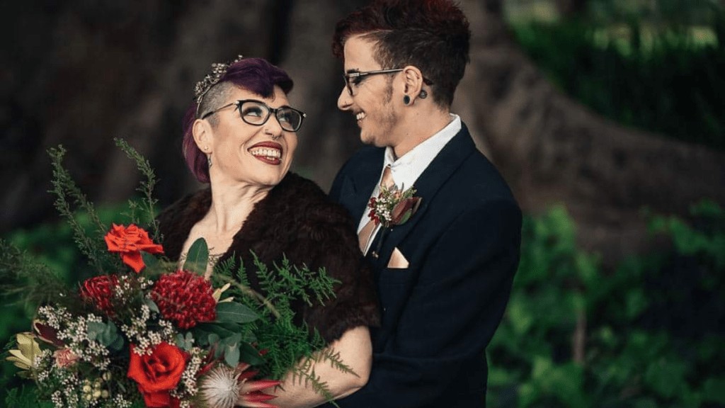 Two people on their wedding day. They are dressed in black and holding a red bunch of flowers whilst smiling at each other.