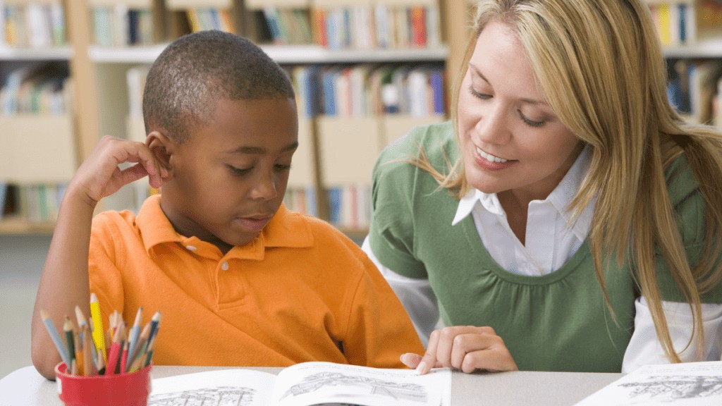 A teacher with long hair smiles at her young male student as she points at the open workbook in front of him on the table. The student is touching their face and looking interested.