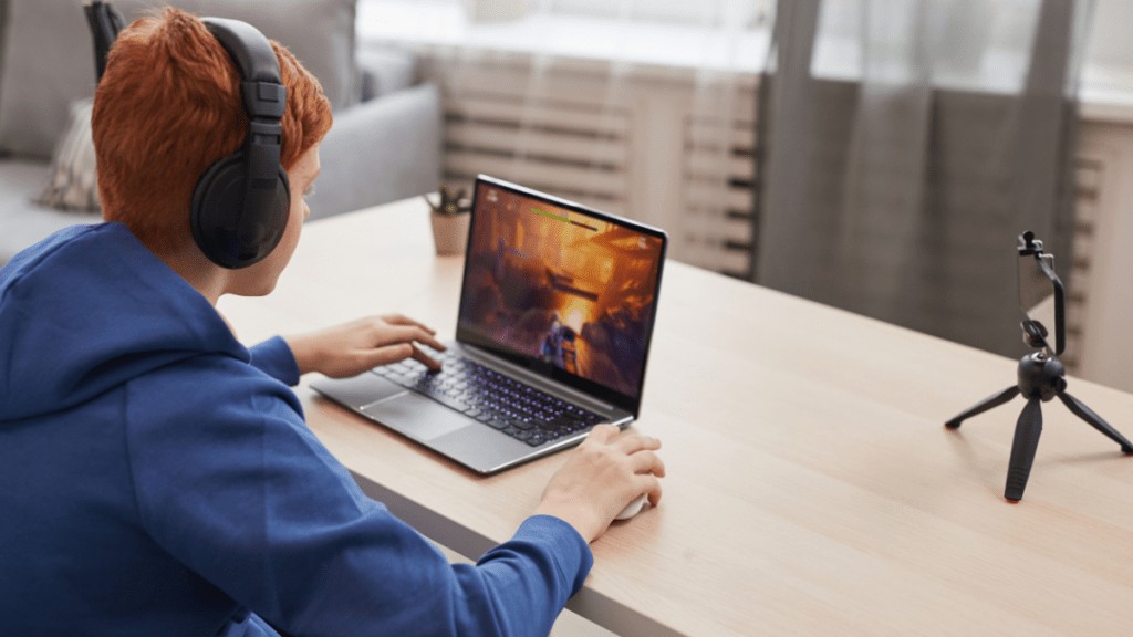 A young boy with red hair is playing a video game on his laptop. He is wearing headphones and there is a camera pointed at him for streaming.