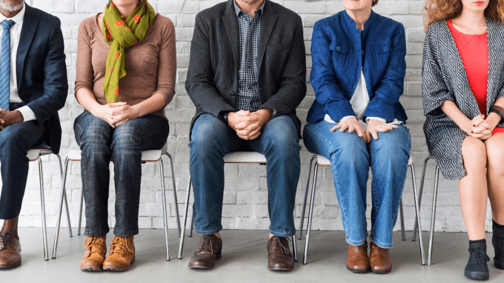 A line up of five people dressed professionally. All are sitting with their hands clasped neatly. They are sitting in front of a grey brick wall, their faces are not visible.
