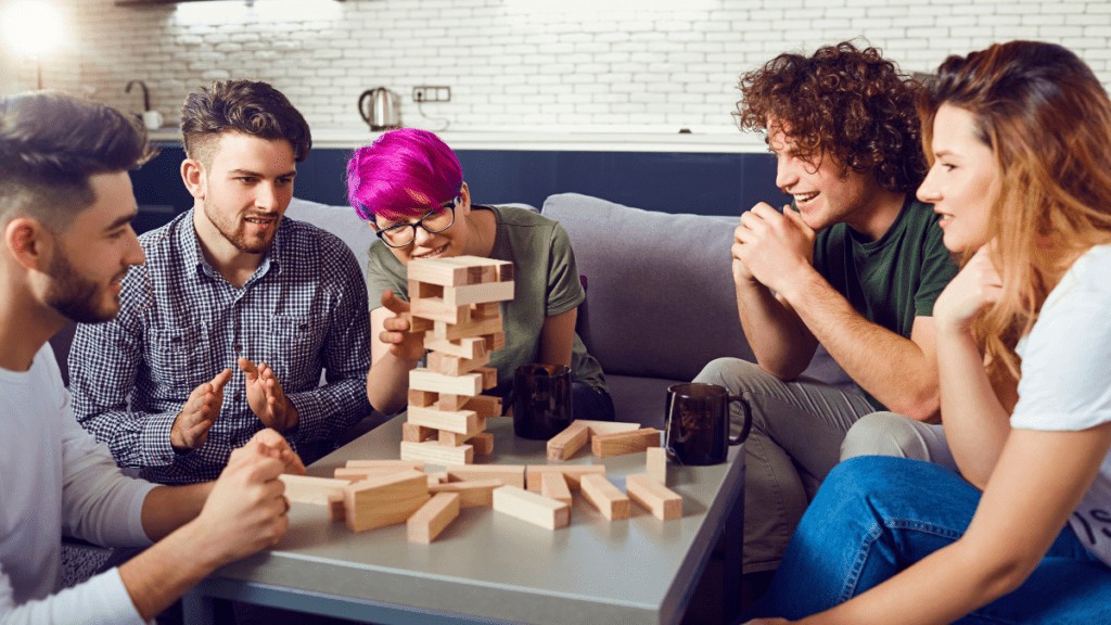 A diverse group of friends sit at a coffee table, playing Jenga together. All are smiling.
