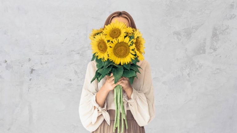 A woman holds a bunch of sunflowers in front of her face, completely covering it. She is standing in front of a grey backdrop.