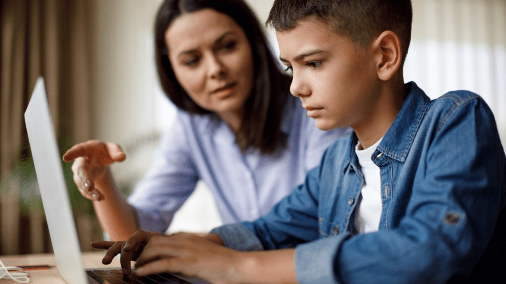 A mother and son are at a laptop. The son is using the laptop and the mother is pointing at the screen. Both look serious.