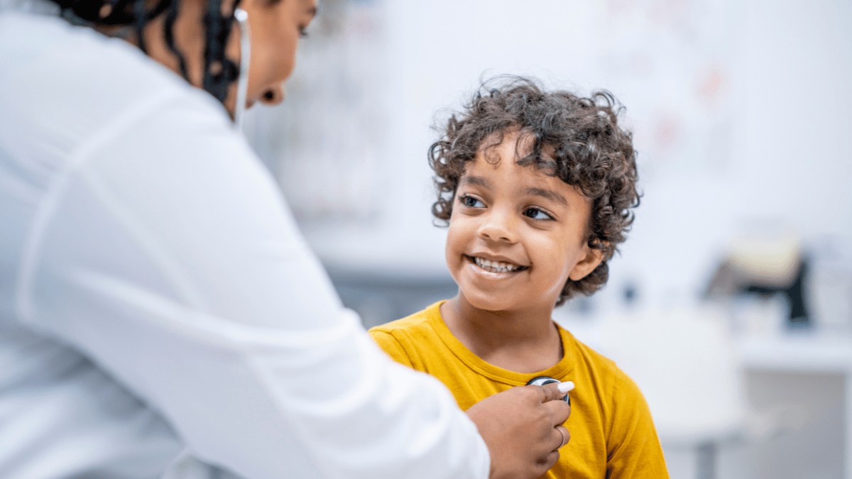 A young boy of colour is getting a physical check up. A doctor has her back to the camera and is holding a stethoscope to the boy's chest. The boy is smiling at the doctor.