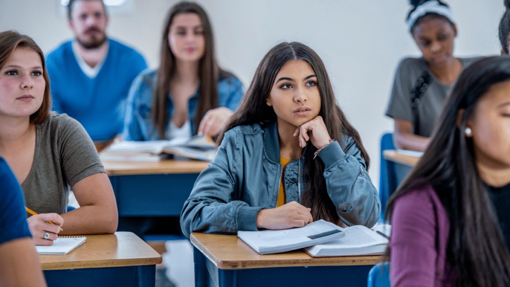 A group of high school students look attentively forward at their teacher. The focus is on one female student with long dark hair and a blue jacket, who has her chin rested on her hand.