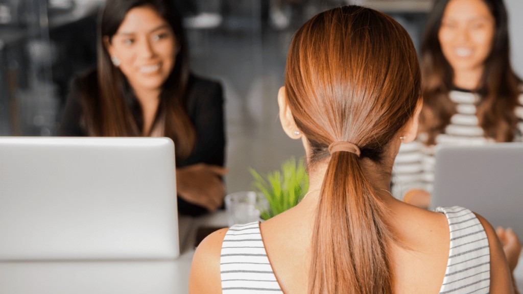 The back of a woman's head, only her ponytail can be seen. She is facing two woman who are smiling and sitting behind laptop screens.