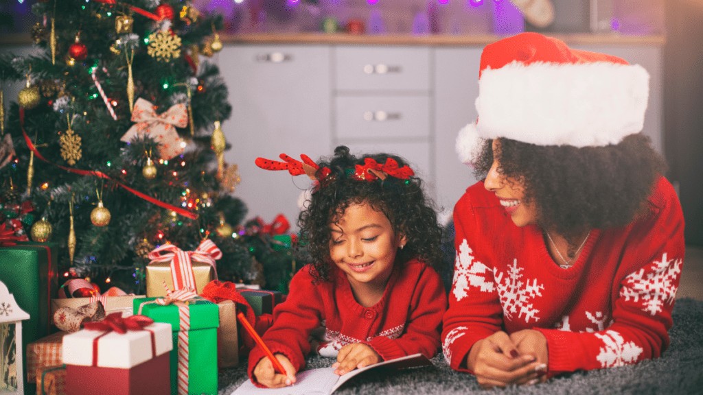A mother and daughter are dressed in Santa hats and festive sweaters. They are laying down in front of the Christmas tree and gifts. The daughter is writing a list on pencil and paper and the mother is smiling at her.