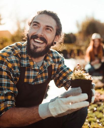 A young man with a beard is working at a nursery. He is holding a seedling, crouched, and smiling away from the camera.