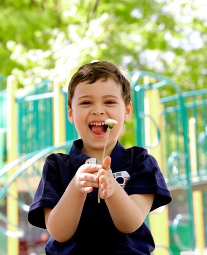 A young boy is on the playground. He is holding a dandelion and laughing joyously at the camera.