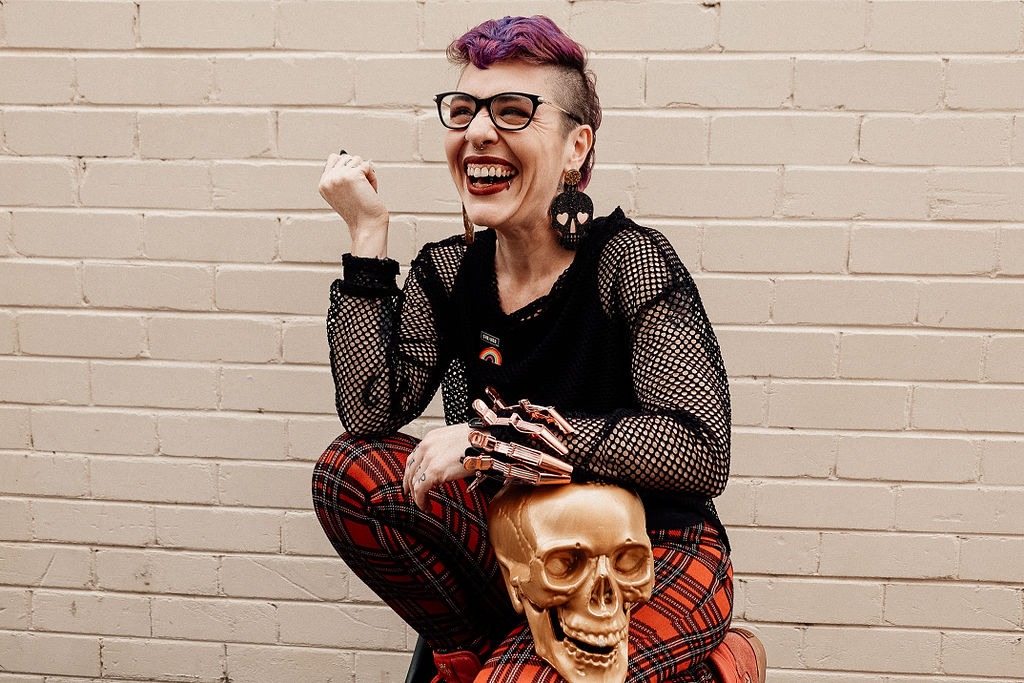 A person with purple and shaved hair is wearing glasses, a mesh top and tartan pants. They are crouching next to a golden skull shape and smiling.