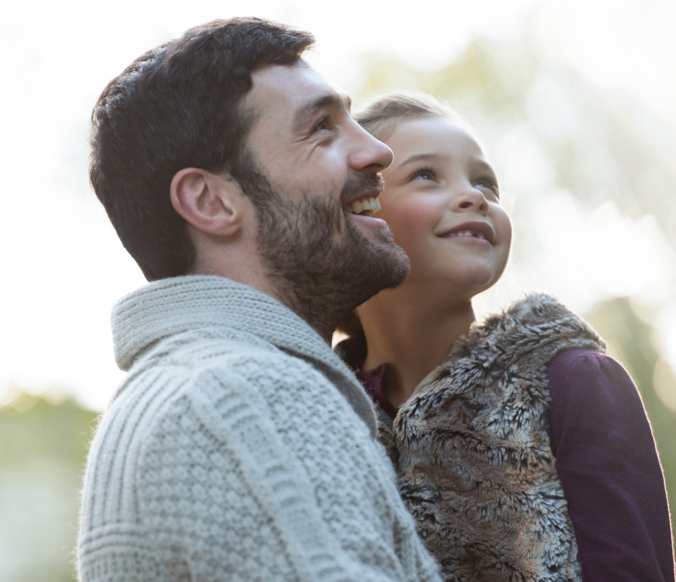 A young father wearing a sweater is holding his daughter in his arms. They are both looking to the right at the sky and smiling.