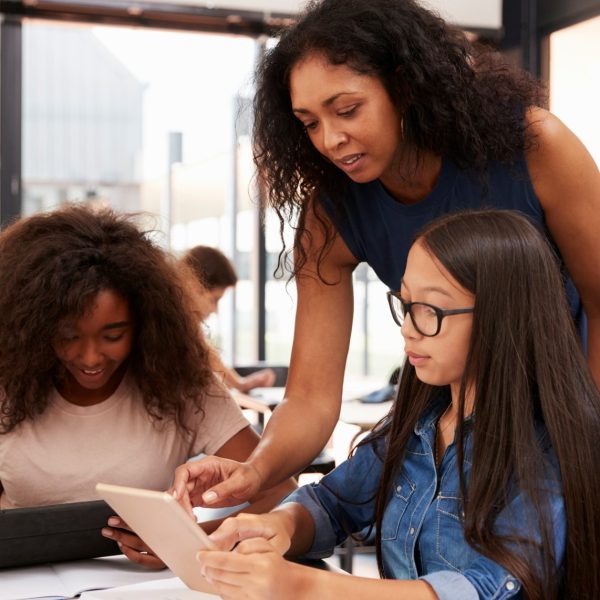 A woman of colour is leaning over two female students. The student on the left is a girl of colour, smiling at an iPad. The student on the right is an Asian girl with long hair and glasses, also looking at an iPad.