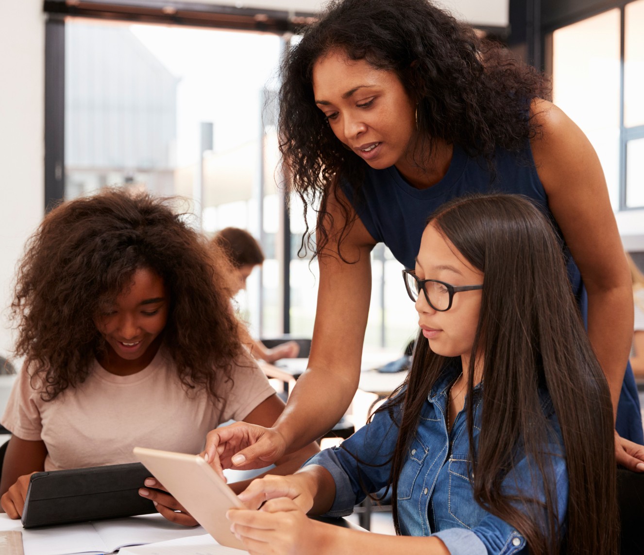 A woman of colour is leaning over two female students. The student on the left is a girl of colour, smiling at an iPad. The student on the right is an Asian girl with long hair and glasses, also looking at an iPad.