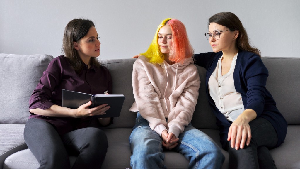 A mother sits with her daughter on a couch. The daughter is of teenage years, and has yellow and orange split dyed hair. The mother and daughter are listening to a woman sitting next to the daughter, who is dressed professionally and writing in an open notebook.