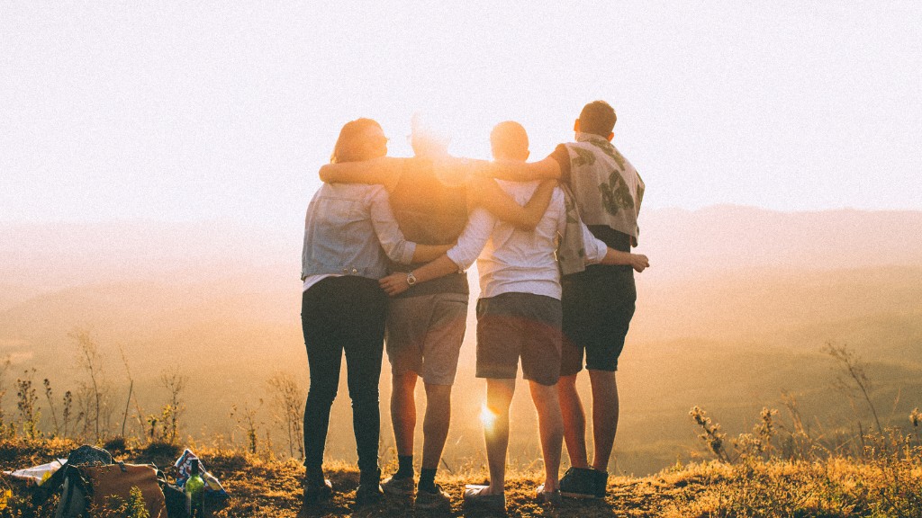 A group of four friends stand with their backs to the camera. They have their arms around each other and are standing on a mountain landscape, across a sunset sky.