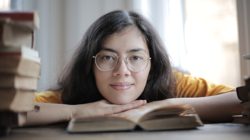 A girl wearing glasses has a pile of books next to her. She is sitting at a table and leaning her hand and chin on an open book. She is smiling softly.