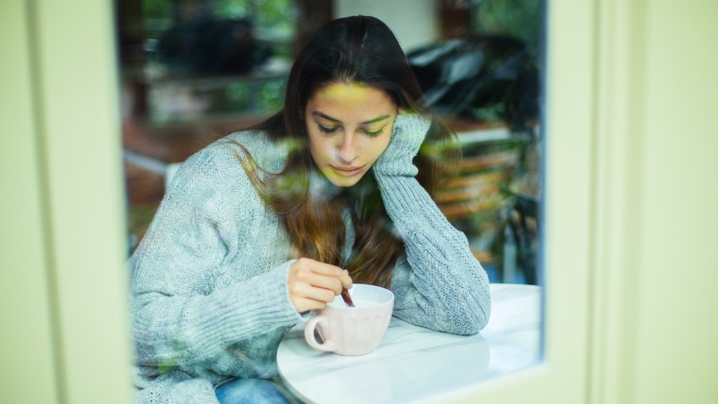 A woman sits inside, viewed through a window. She is stirring her coffee and looking pensively, holding her head in her hand.