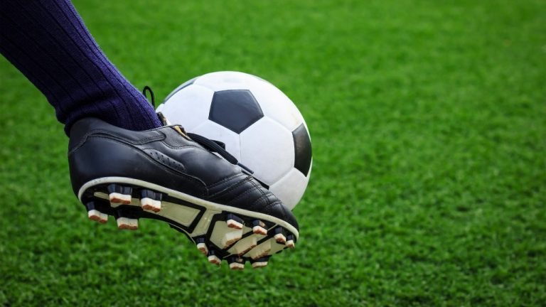 A grass background and a close up of a foot kicking a soccer ball.