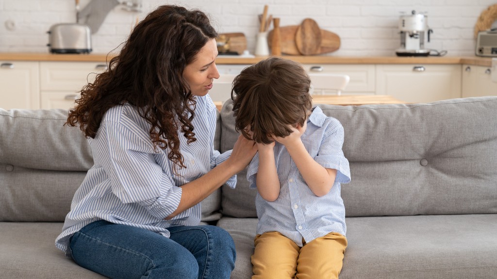 A mother sitting on a couch comforts her young son, who has his face buried in tears in his hands.