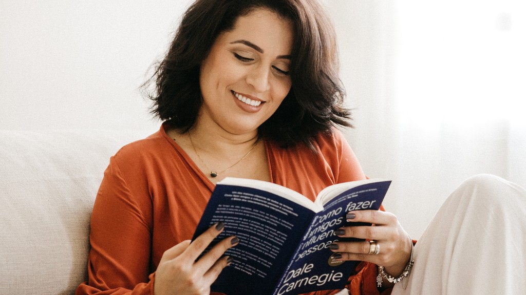 A woman with dark hair is reading a book while sitting on a sofa. She is smiling as she reads.
