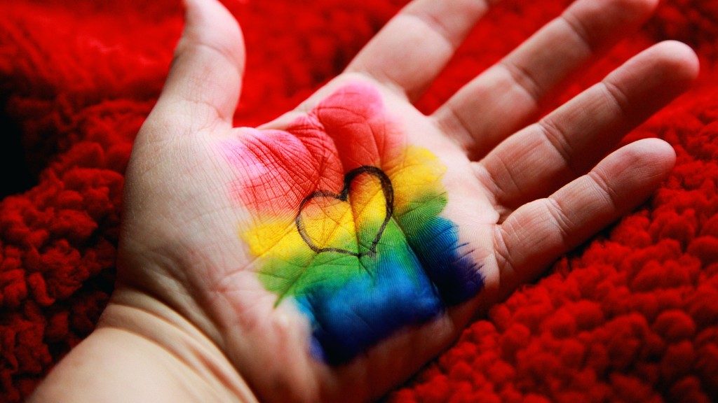 A child's hand on a red rug. There is a rainbow flag and a black heart outline painted onto their palm, and their palm is facing up towards the camera.